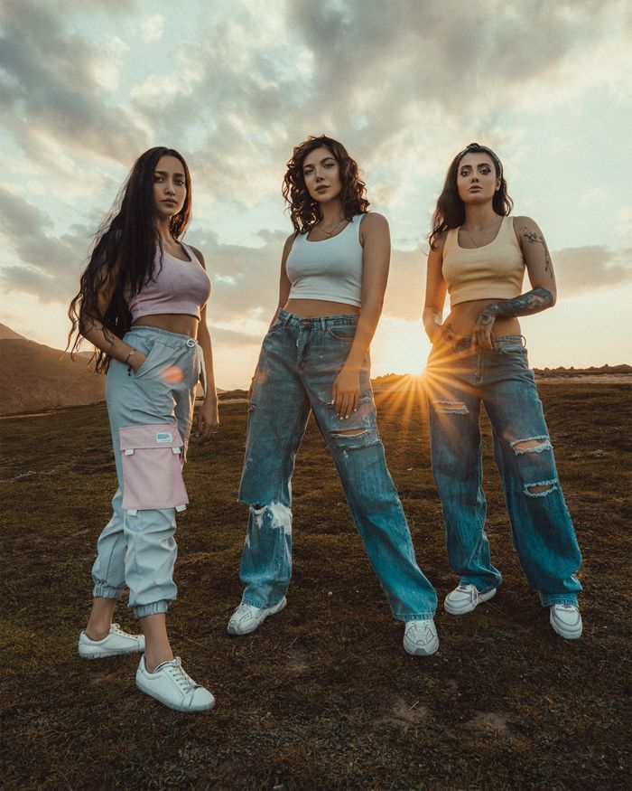 Full body of charming girlfriends in trendy clothes standing in grassy field on hill at sunset